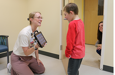 Image of a provide, a white female with blonde hair, holding a touch screen up for a young white child to look at, behind them a white female with dark hair crouches down and smiles through the door.